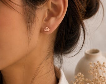 May include: A small, rose gold-toned stud earring shaped like a stylized hedgehog. The earring features a smooth, flat surface with a simple outline and small, evenly spaced cutouts along the back to represent the quills of the animal.