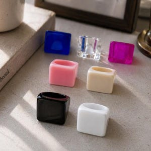 May include: A collection of square-shaped rings in various colors, including blue, pink, clear, white, black, and cream. The rings are arranged on a light gray surface, with a book and a framed picture in the background.
