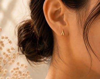 May include: A small gold stud earring shaped like the Eiffel Tower. The earring features a metallic finish and a detailed silhouette of the structure, securely fastened to the earlobe. The design is simple, compact, and polished in appearance.