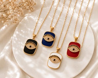 May include: Gold-toned necklaces with rectangular pendants in black, blue, white, and red. Each pendant features an eye design with a central black gem and surrounding clear gems. The necklaces are displayed on a white plate.