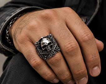 May include: A silver-toned ring featuring a central skull motif set against a shield-like background with crossed bands and small stud details. The metal has a polished finish on the skull and a darkened, textured finish on the surrounding elements.