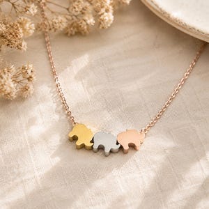 May include: A delicate necklace featuring three small elephant-shaped charms suspended from a thin chain. The charms are finished in three distinct metallic tones: a yellow gold, a silver, and a rose gold, creating a tri-color effect on the chain.