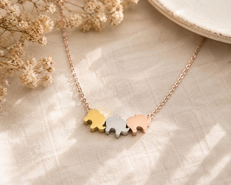 May include: A delicate necklace featuring three small elephant-shaped charms suspended from a thin chain. The charms are finished in three distinct metallic tones: a yellow gold, a silver, and a rose gold, creating a tri-color effect on the chain.