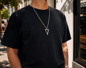 May include: A silver necklace with a triangular pendant hangs against a black t-shirt. The pendant is a hollow triangle. The necklace chain is a series of small, connected links. The person is wearing a silver bracelet.