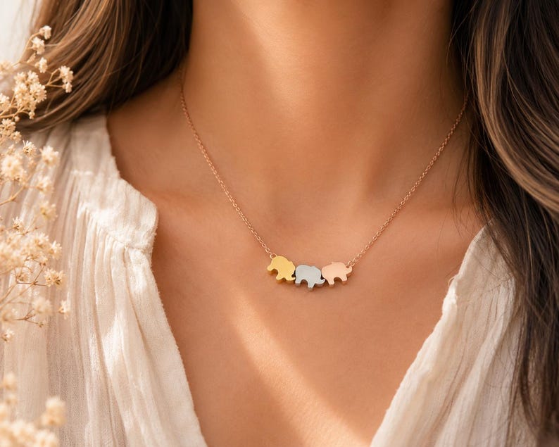 May include: A delicate necklace featuring three small elephant-shaped charms suspended from a thin chain. The charms are finished in three distinct metallic tones: a yellow gold, a silver, and a rose gold, arranged horizontally in a row.