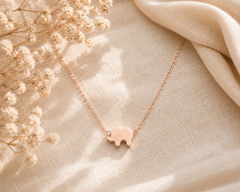 May include: A rose gold-toned necklace featuring a small, flat elephant-shaped pendant centered on a delicate cable chain. The metal has a smooth, polished finish, and the design is simple, showcasing the silhouette of an elephant in profile.