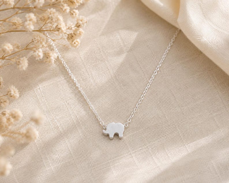 May include: A silver-toned necklace featuring a small, flat elephant-shaped pendant centered on a delicate, thin link chain. The pendant has a smooth, solid surface and is attached directly to the chain on both sides of the elephant figure.