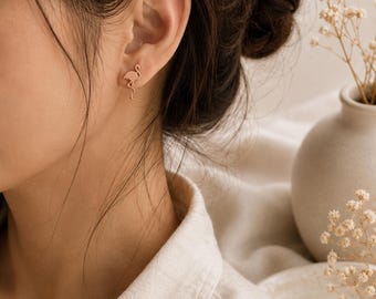 May include: A small, rose gold-toned stud earring shaped like a flamingo in a standing position. The earring features a minimalist silhouette of the bird with a long neck, curved body, and thin legs, securely fastened to the earlobe with a post back.