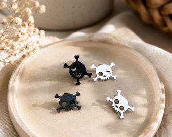 May include: Four stud earrings shaped like skulls with crossed bones behind them. Two of the earrings are matte black, and two are polished silver. Each earring features a detailed skull design with hollow eye sockets and a jagged jawline.