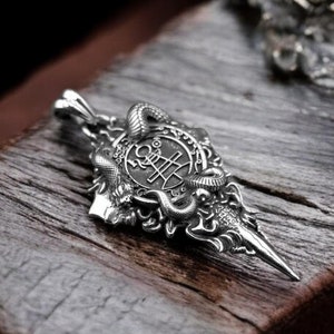 May include: Silver pendant with a detailed design featuring a circular emblem with a symbol and two snakes. The pendant is shaped like a spearhead and has a loop for attaching a chain.