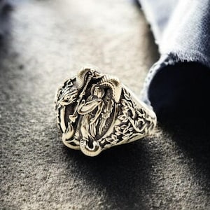 Op de afbeelding: Een zilveren ring met een ingewikkeld ontwerp met een engel met vleugels. De ring ligt op een donkerblauwe stoffen achtergrond.
