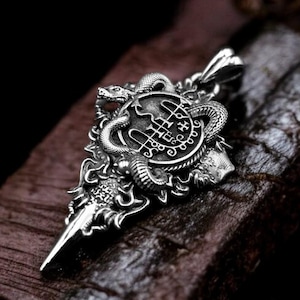 Puede incluir: Un colgante de plata con un diseño detallado de una daga con una serpiente envuelta alrededor. La cabeza de la serpiente está levantada y la daga tiene una punta puntiaguda. El colgante está sobre una superficie de madera marrón oscura.