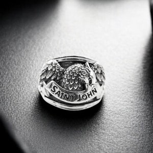 Könnte beinhalten: Ein silberner Ring mit einem detaillierten Design eines Adlers mit ausgebreiteten Flügeln und der Inschrift "SAINT JOHN" um den Adler herum.