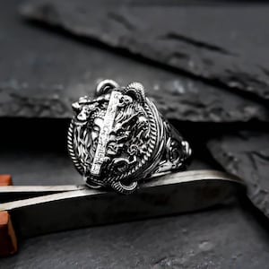 Peut inclure: Une bague en argent avec un design détaillé représentant une tête de dragon et des motifs complexes. La bague est placée sur un fond sombre.