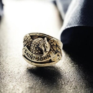 Könnte beinhalten: Ein goldener Ring mit einem geflügelten Adler-Design und der Inschrift "SAINT JOHN" auf der Band.