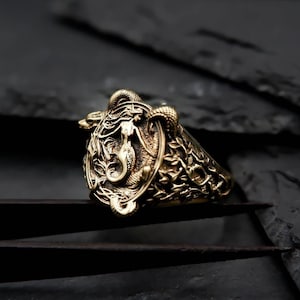 May include: A gold-toned ring with a detailed design featuring a stylized animal head and intricate patterns. The ring is set against a dark background.