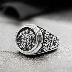 Könnte beinhalten: Ein silberner Ring mit einem detaillierten Design, das eine Schildkröte auf der Vorderseite des Rings zeigt.