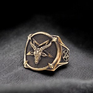 May include: A gold-toned ring with a black pentagram and a goat's head in the center. The ring is set on a black fabric background.