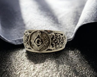 May include: A silver ring with a detailed design featuring a Masonic symbol, a compass and square, with the letter "G" in the center.