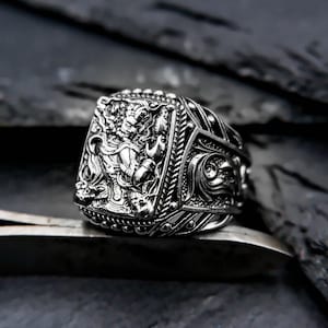 Peut inclure: Une bague en argent avec un motif complexe représentant une créature mythique. La bague est placée sur un fond sombre.