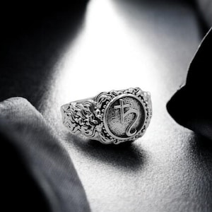 May include: A silver ring with an ornate design featuring a symbol of a cross and a crescent moon.