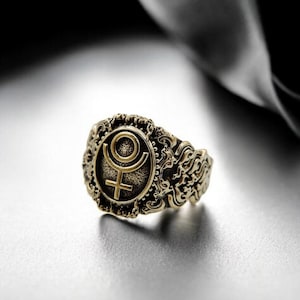 May include: A gold ring with an ornate design featuring a symbol of a circle with a cross inside. The ring is on a white surface.