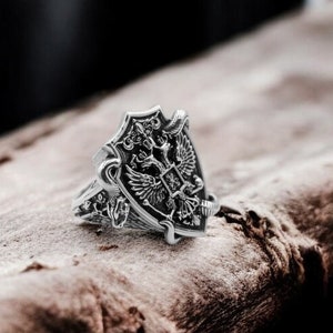 Puede incluir: Un anillo de plata con un diseño detallado de un escudo con dos águilas y otros símbolos.