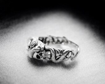 May include: A silver ring with a detailed design of a skull and snakes. The ring is on a gray fabric background.