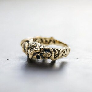 Op de afbeelding: Een goudkleurige ring met een schedel- en slangenontwerp. De ring heeft ingewikkelde details, met de slang die zich om de schedel wikkelt. De ring staat tegen een neutrale achtergrond, wat het vakmanschap van de sieraden benadrukt.
