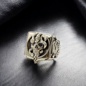 Op de afbeelding: Een zilveren ring met een vierkant gezicht met een gedetailleerd schedelontwerp met een slang eromheen gewikkeld. De ring heeft een getextureerde band.