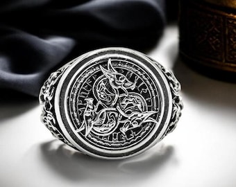 May include: A silver-toned ring featuring a circular face with an embossed design of three intertwined dragon-like creatures. The metal surface has a dark, oxidized finish in the recessed areas to highlight the intricate details of the relief.