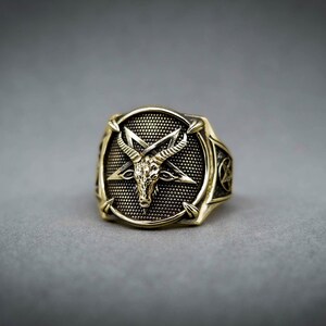 May include: A gold-toned ring with a pentagram design featuring a goat's head in the center. The ring has a textured surface and a detailed design.