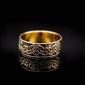 Könnte beinhalten: Ein goldfarbener Ring mit einem aufwendigen keltischen Knotenmuster. Der Ring wird auf einer reflektierenden schwarzen Oberfläche präsentiert, die das detaillierte Muster und den metallischen Glanz des Rings hervorhebt. Das Design zeigt verschlungene Muster.
