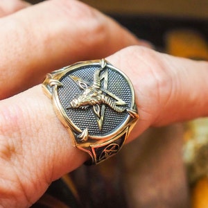 May include: A gold-toned ring with a black pentagram and a goat's head in the center. The ring is worn on a finger.