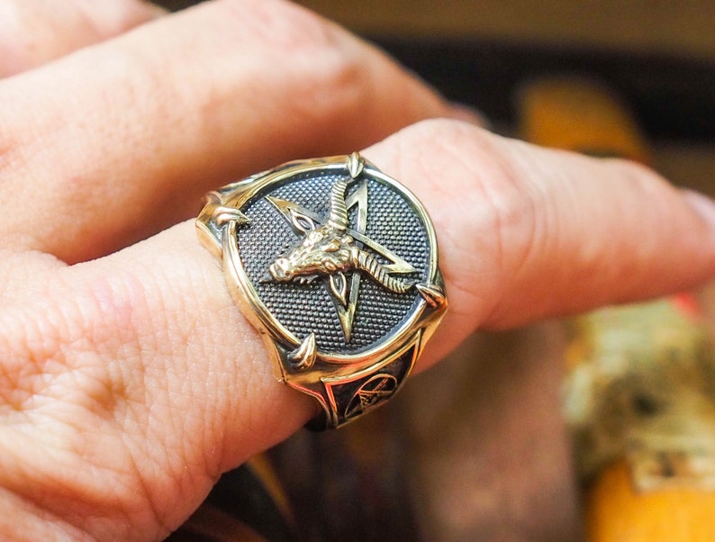 May include: A gold-toned ring with a black pentagram and a goat's head in the center. The ring is worn on a finger.