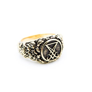 May include: A gold-coloured ring with an embossed design. The ring features a central emblem within a circular frame, surrounded by an ornate, swirling pattern. The emblem appears to be a stylized symbol. The ring is set against a white background.