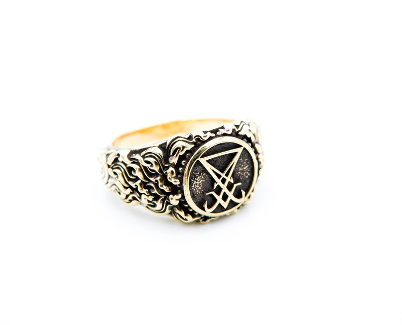May include: A gold-coloured ring with an embossed design. The ring features a central emblem within a circular frame, surrounded by an ornate, swirling pattern. The emblem appears to be a stylized symbol. The ring is set against a white background.