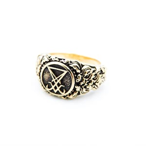 May include: A gold-toned ring with an elaborate design. The ring showcases a central symbol within a circular frame, encircled by swirling patterns. The ring is set against a white backdrop, accentuating its intricate details.