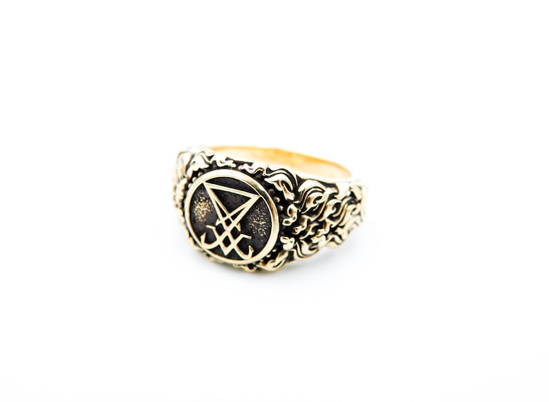 May include: A gold-toned ring with an elaborate design. The ring showcases a central symbol within a circular frame, encircled by swirling patterns. The ring is set against a white backdrop, accentuating its intricate details.
