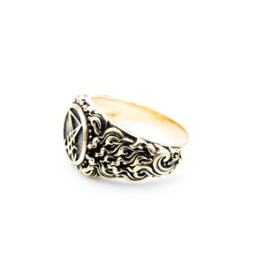May include: Gold-coloured ring with an intricate design. The ring has an oval face with a pentagram symbol. The band features a swirling, flame-like pattern. The ring is set against a white background.