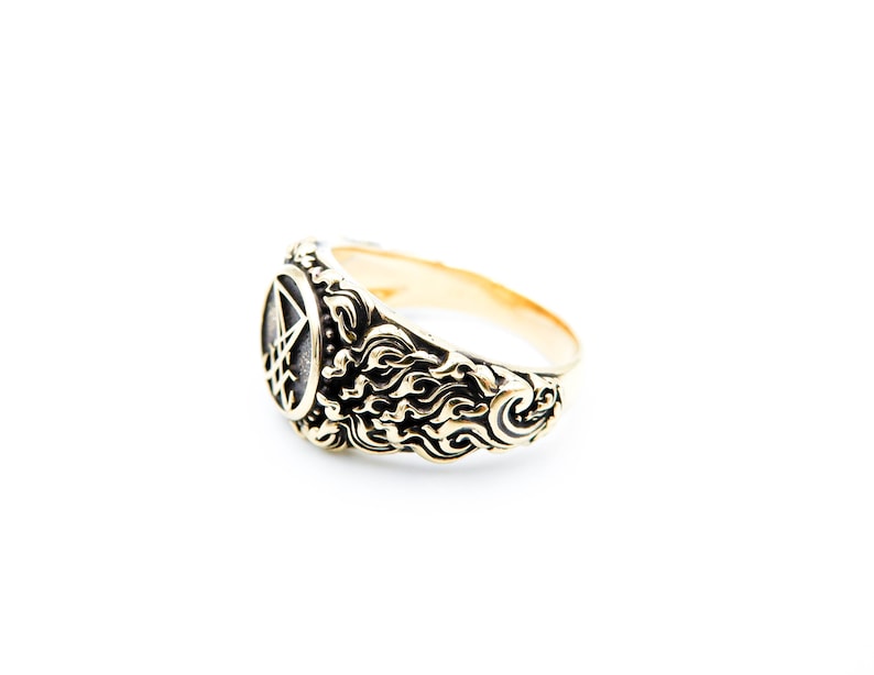 May include: Gold-coloured ring with an intricate design. The ring has an oval face with a pentagram symbol. The band features a swirling, flame-like pattern. The ring is set against a white background.