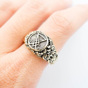 May include: An antique-style ring with a circular emblem featuring a stylized symbol. The ring has an ornate, textured band with a gold-tone finish. The emblem is set against a darker, textured background.