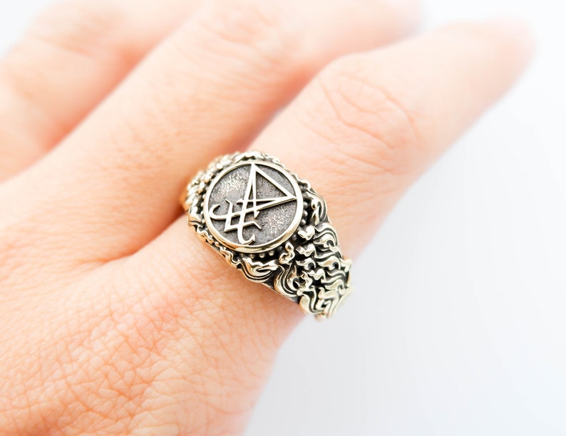 May include: An antique-style ring with a circular emblem featuring a stylized symbol. The ring has an ornate, textured band with a gold-tone finish. The emblem is set against a darker, textured background.