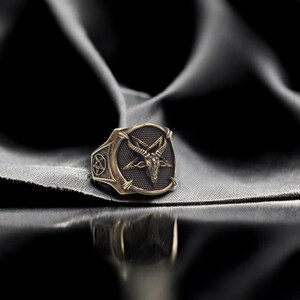 May include: A gold-toned ring with a black pentagram and a goat's head design. The ring is on a dark gray fabric background.