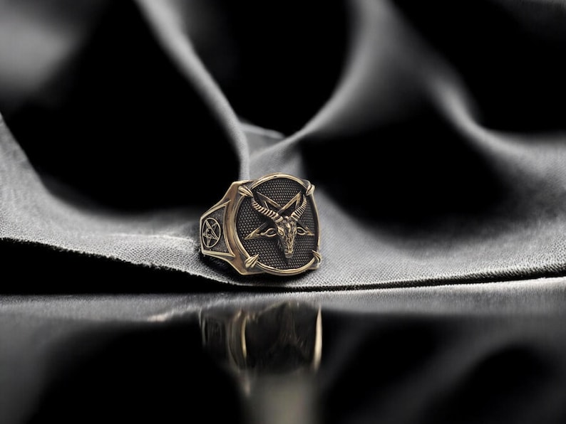 May include: A gold-toned ring with a black pentagram and a goat's head design. The ring is on a dark gray fabric background.