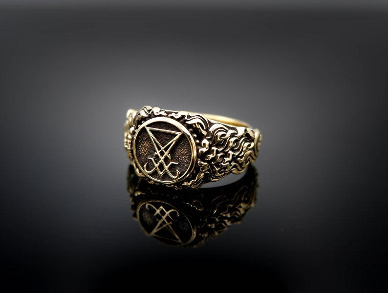 May include: A gold ring with an oval emblem displaying a symbol. The ring has an ornate, textured band with a detailed design. The emblem's symbol is a stylized inverted triangle with a cross and other markings.