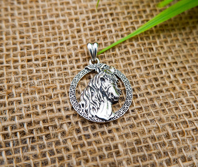 May include: Silver pendant featuring a detailed horse head design with a decorative border. The pendant is on a burlap background.