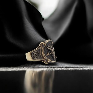 May include: A gold ring with a black and silver design featuring a pentagram and a sword. The ring is on a black fabric background.