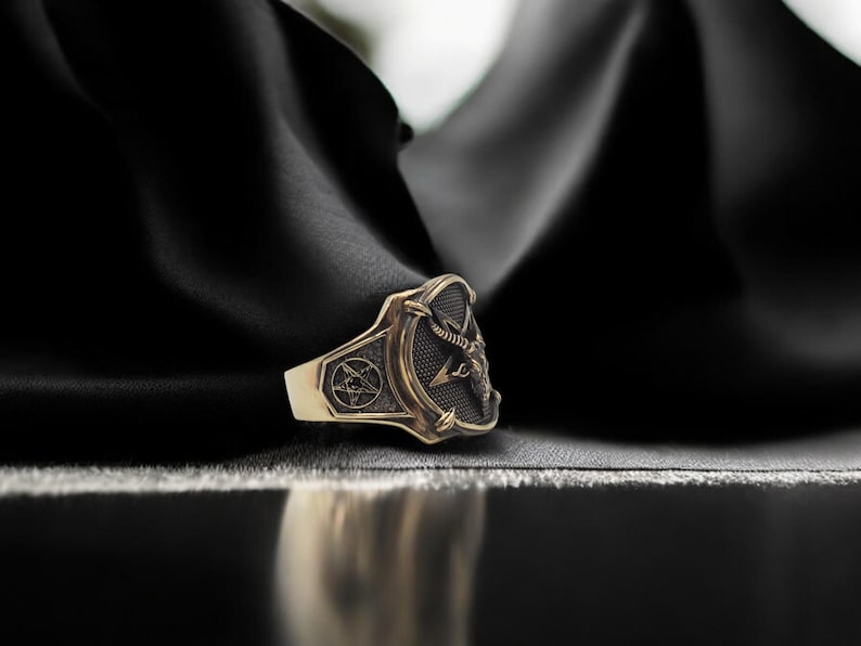 May include: A gold ring with a black and silver design featuring a pentagram and a sword. The ring is on a black fabric background.