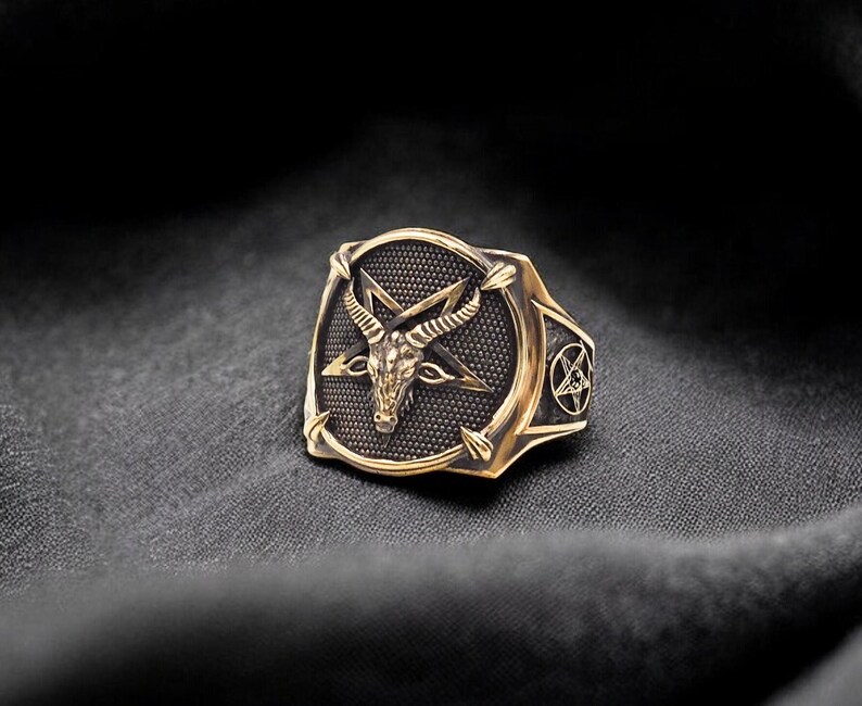 May include: A gold-toned ring with a black pentagram and a goat's head in the center. The ring is set on a black fabric background.